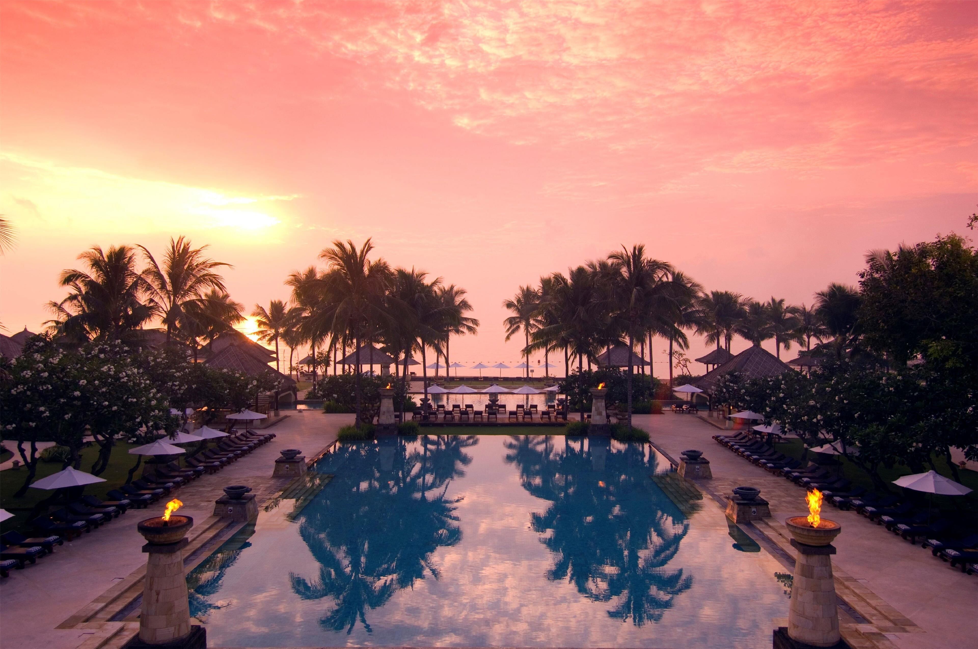 A resort pool with lounge chairs and umbrellas, surrounded by palm trees and umbrellas, reflecting the sunset.