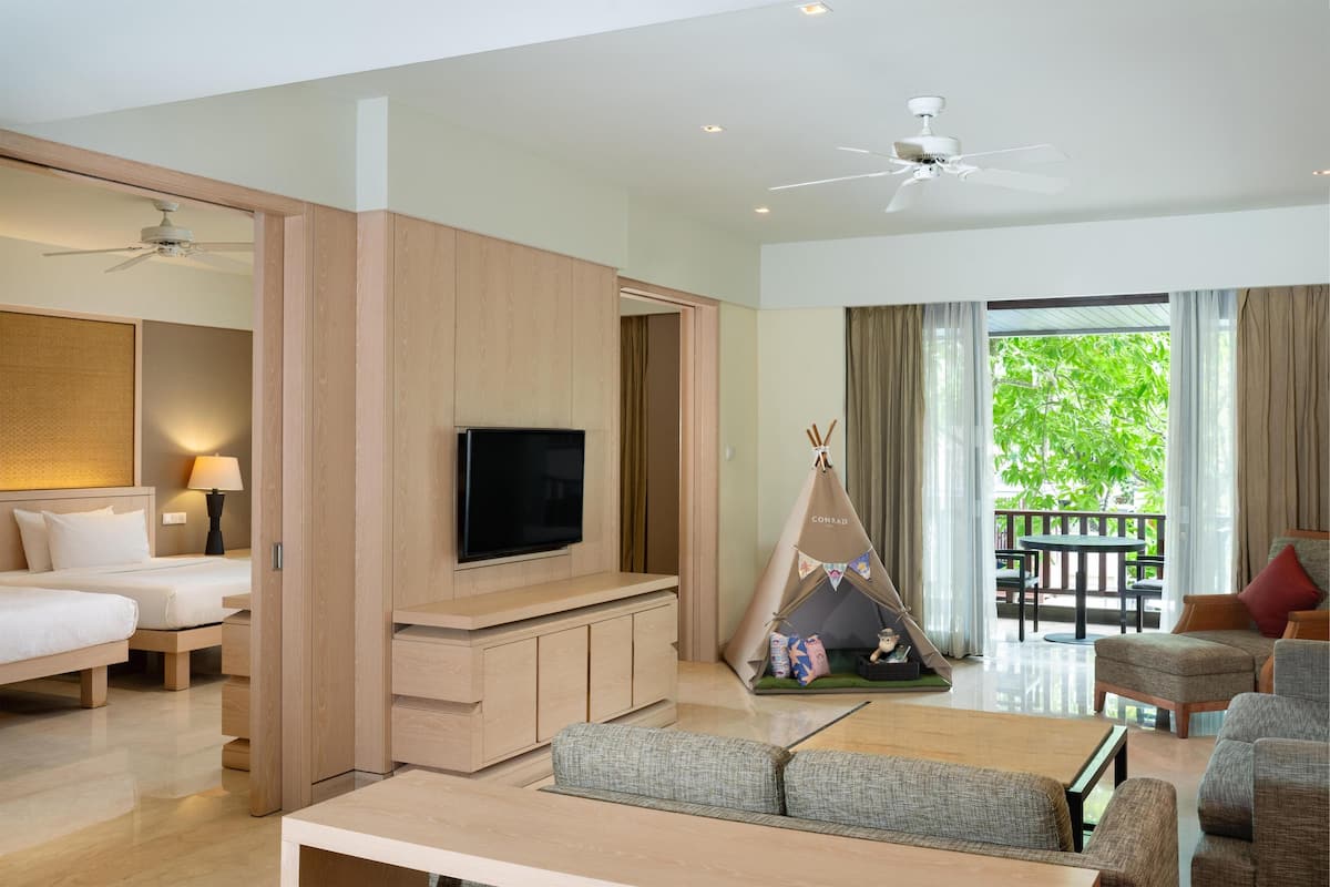Habitación de hotel con televisión, cama, zona de estar con mesa y balcón con vistas a zonas verdes.