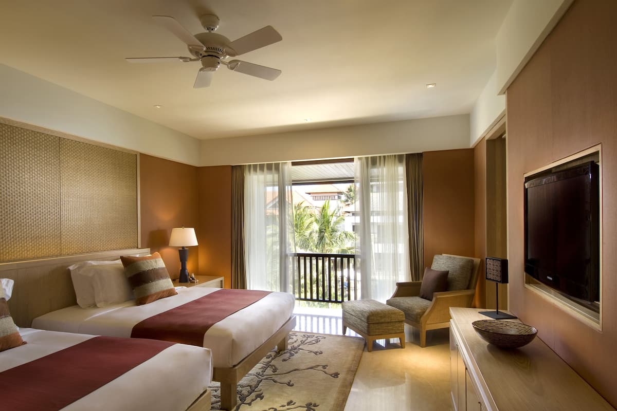Habitación de hotel con dos camas, ventilador de techo, televisión, balcón con cortinas y vista a palmeras.