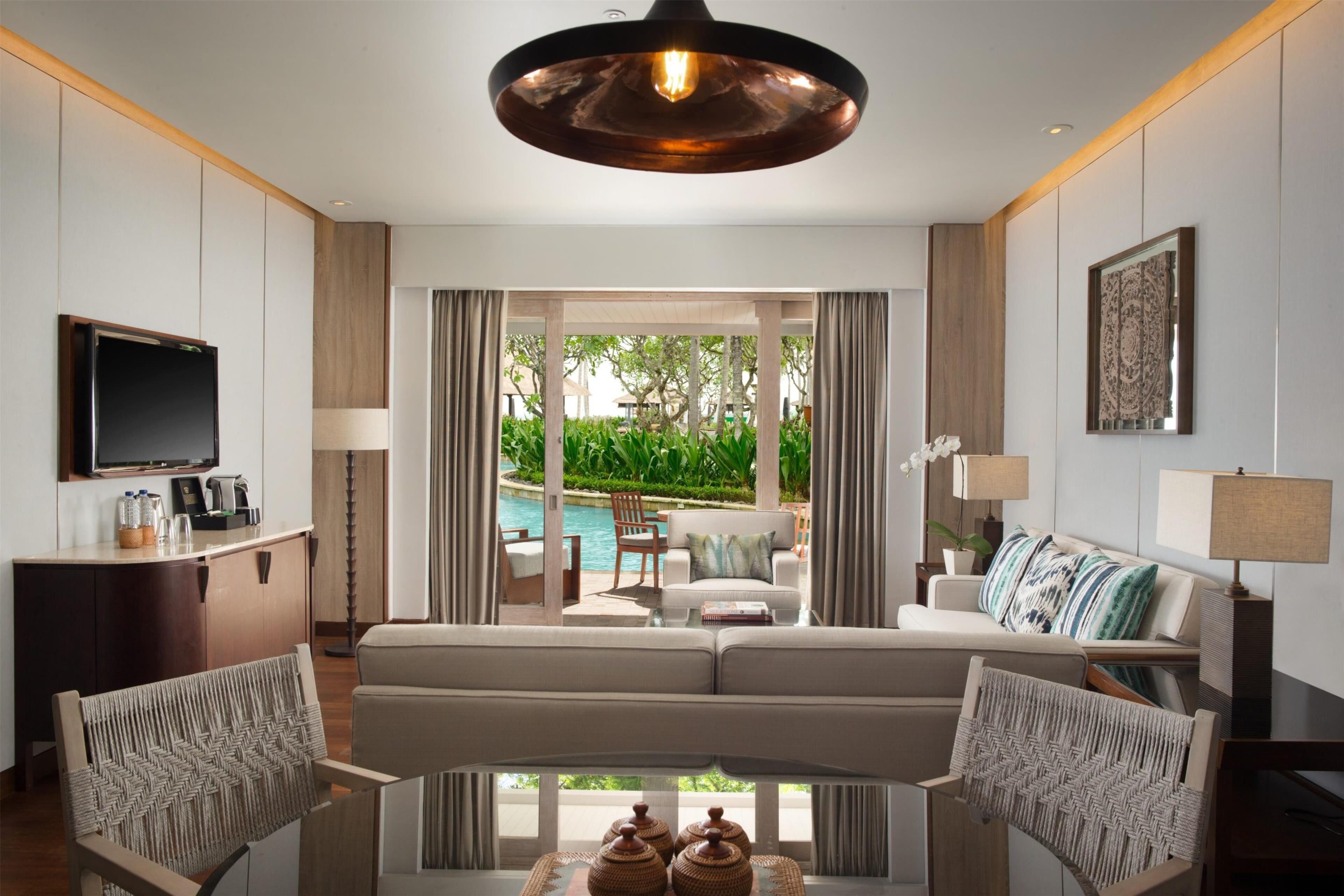 A modern living room with a sofa, armchairs, a coffee table, and a television. There is a view of a pool and greenery outside.