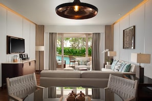 A modern living room with a sofa, armchairs, a coffee table, and a television. There is a view of a pool and greenery outside.
