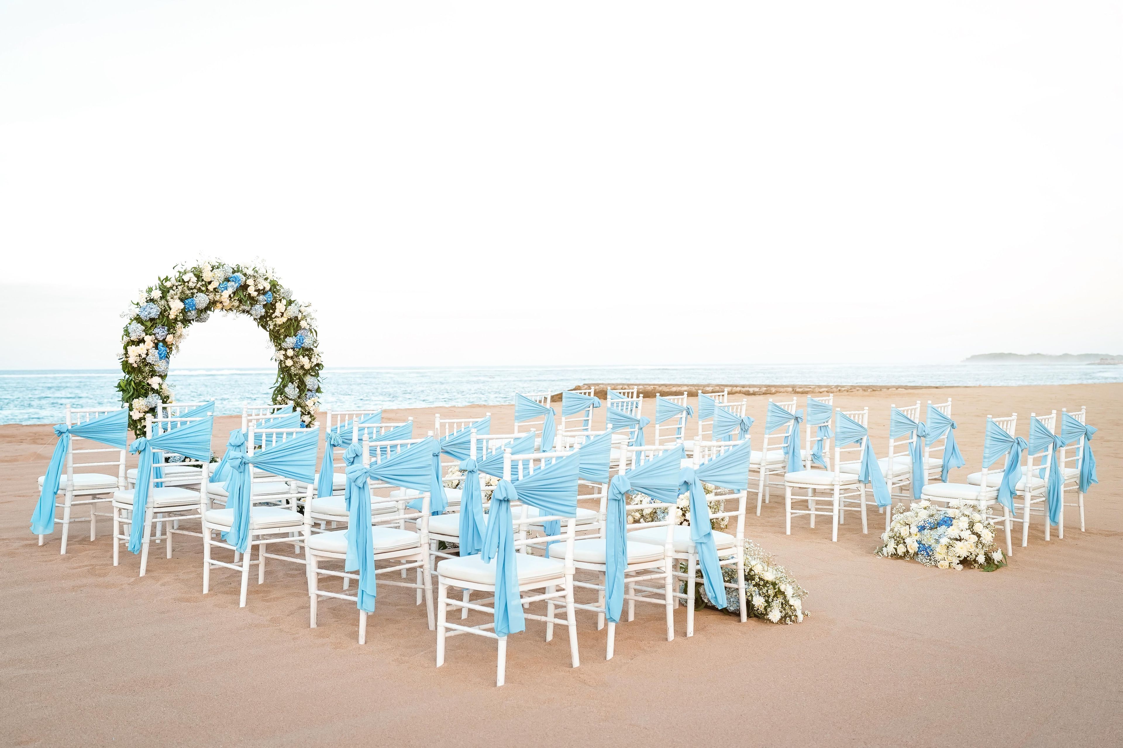 A beach wedding setup with white chairs and a floral arch.