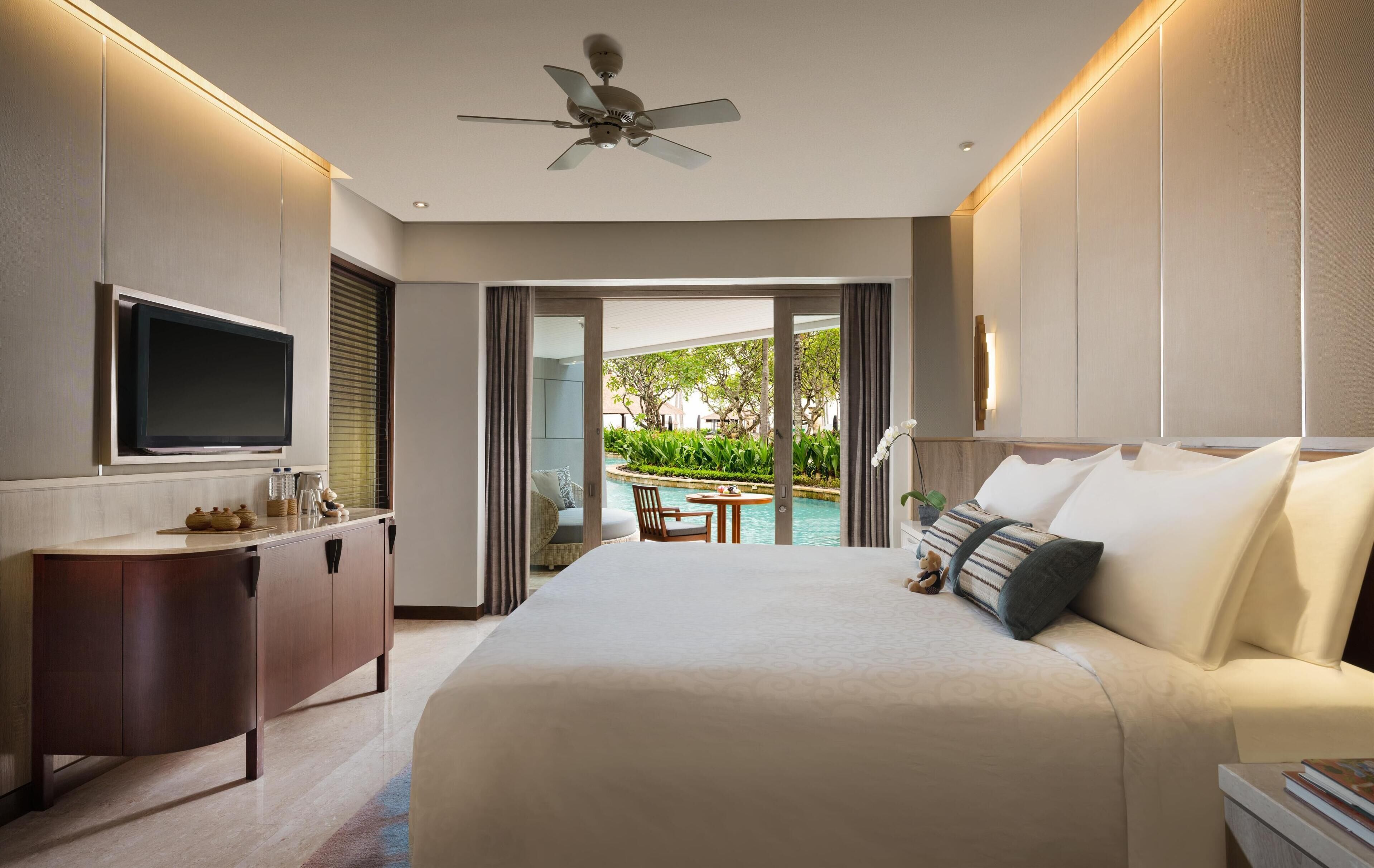 A modern hotel room with a large bed, a TV, a desk, and a view of a pool and greenery.