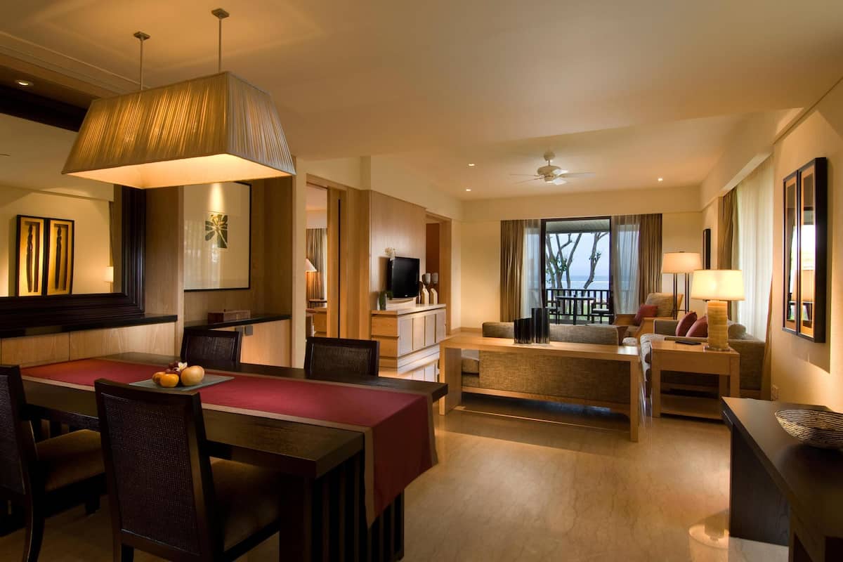 Una habitación de hotel moderna con zona de comedor, sala de estar con sofá y sillones, televisión y un ventanal con vistas.
