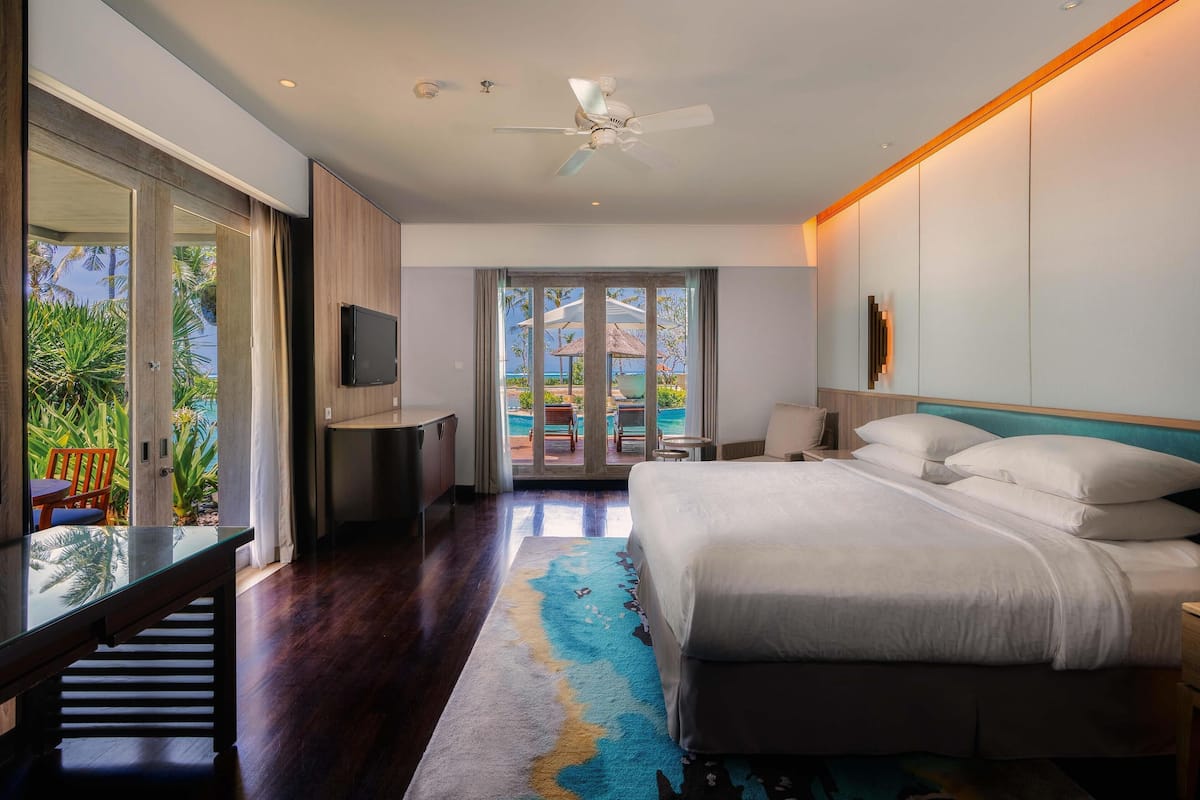Una habitación de hotel moderna con una cama grande, un televisor, un escritorio y vistas a una piscina y palmeras.