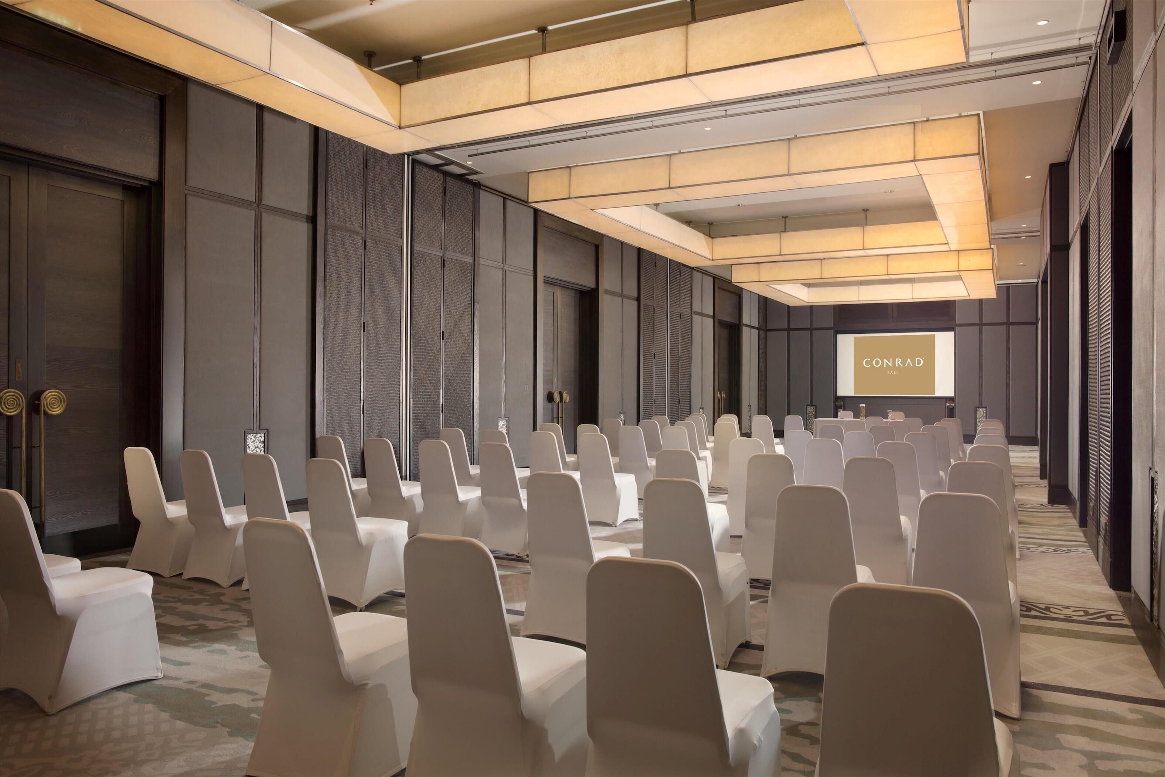 A conference room with rows of white chairs, a large screen displaying 'CONRAD', and a modern ceiling design.