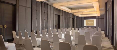 Un salón de conferencias con hileras de sillas blancas, una pantalla grande que muestra 'CONRAD' y un diseño de techo moderno.