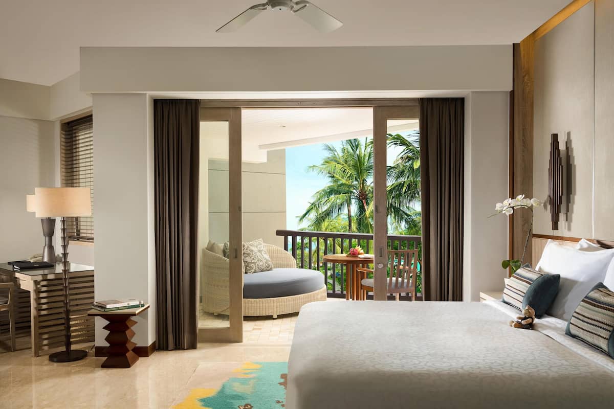 Habitación de hotel con cama, escritorio, silla, balcón y vistas a palmeras, además de ventilador de techo.