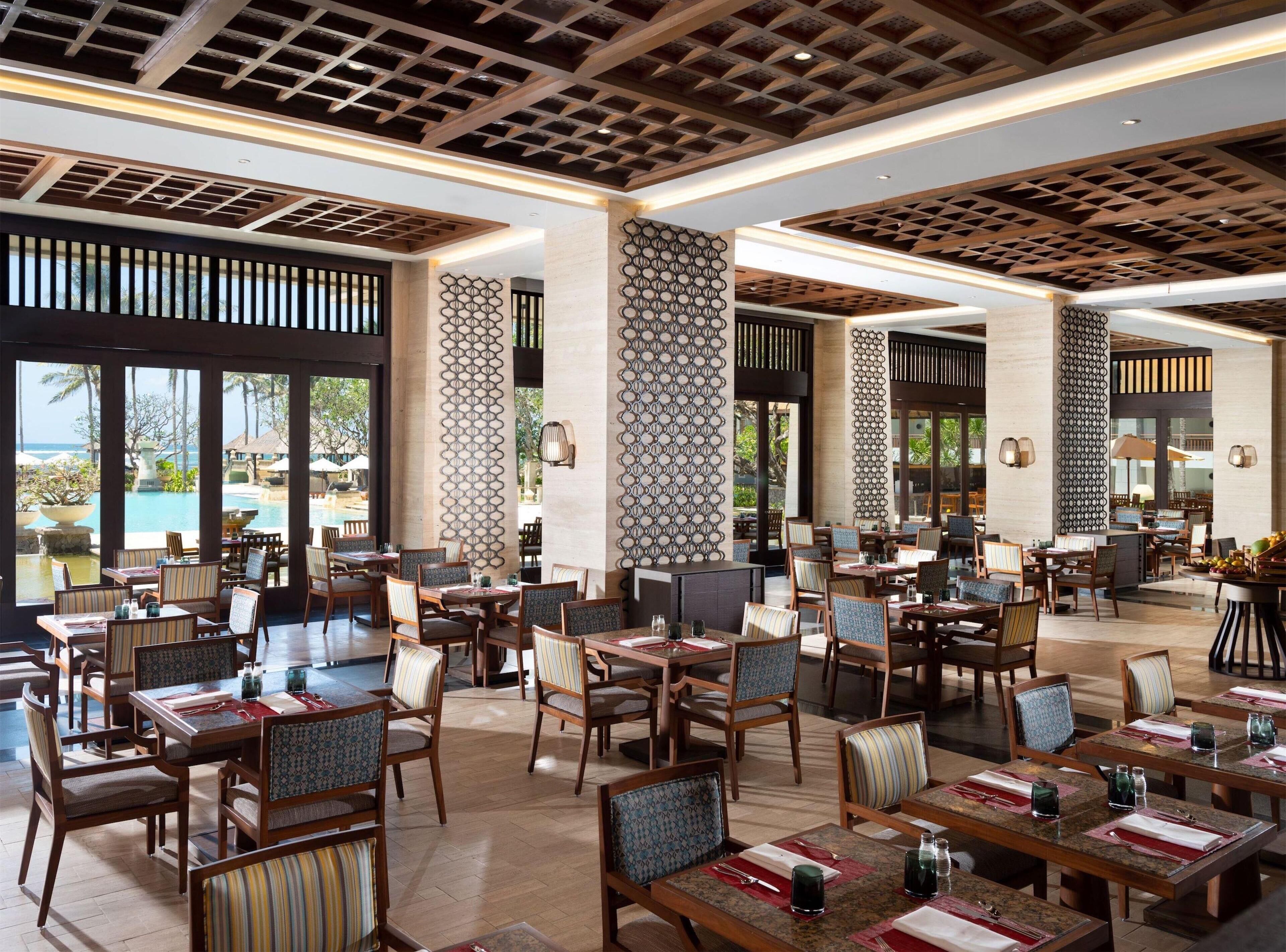 A spacious restaurant with multiple dining tables and chairs, large windows offering a view of the pool and palm trees, and a ceiling with a geometric pattern.