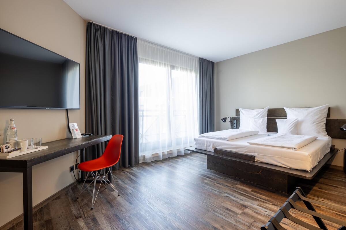A hotel room with a bed, a desk with a chair, a TV, and a window with curtains.
