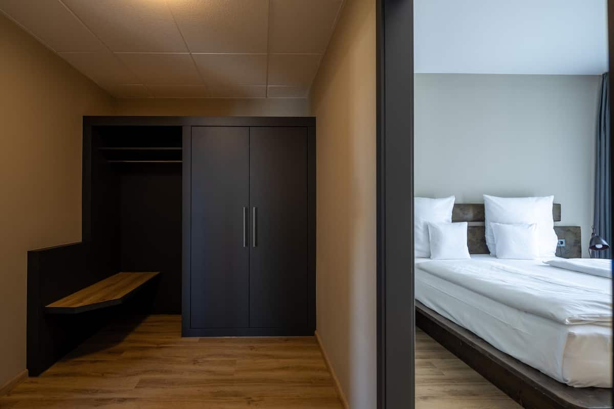 A modern hotel room with a wooden bench, a dark wardrobe, and a neatly made bed.