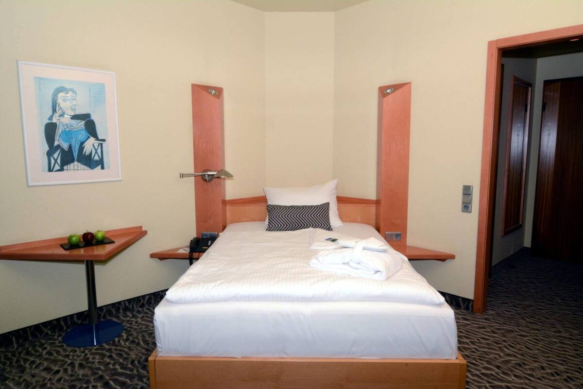 A hotel room with a bed, a small table with fruits, a painting on the wall, and a door leading to another room.