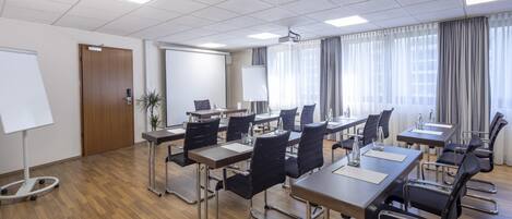Un salón de conferencias con varias mesas y sillas, una pizarra y una pantalla de proyector.