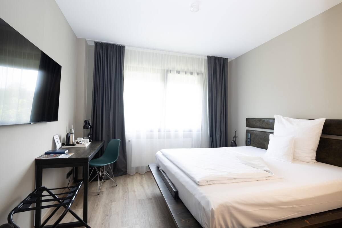 A hotel room with a bed, a desk, a chair, a TV, and a window with curtains.