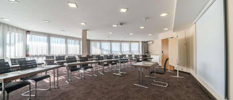 Un salón de conferencias con varias mesas y sillas, ventanas amplias con cortinas y una pizarra.