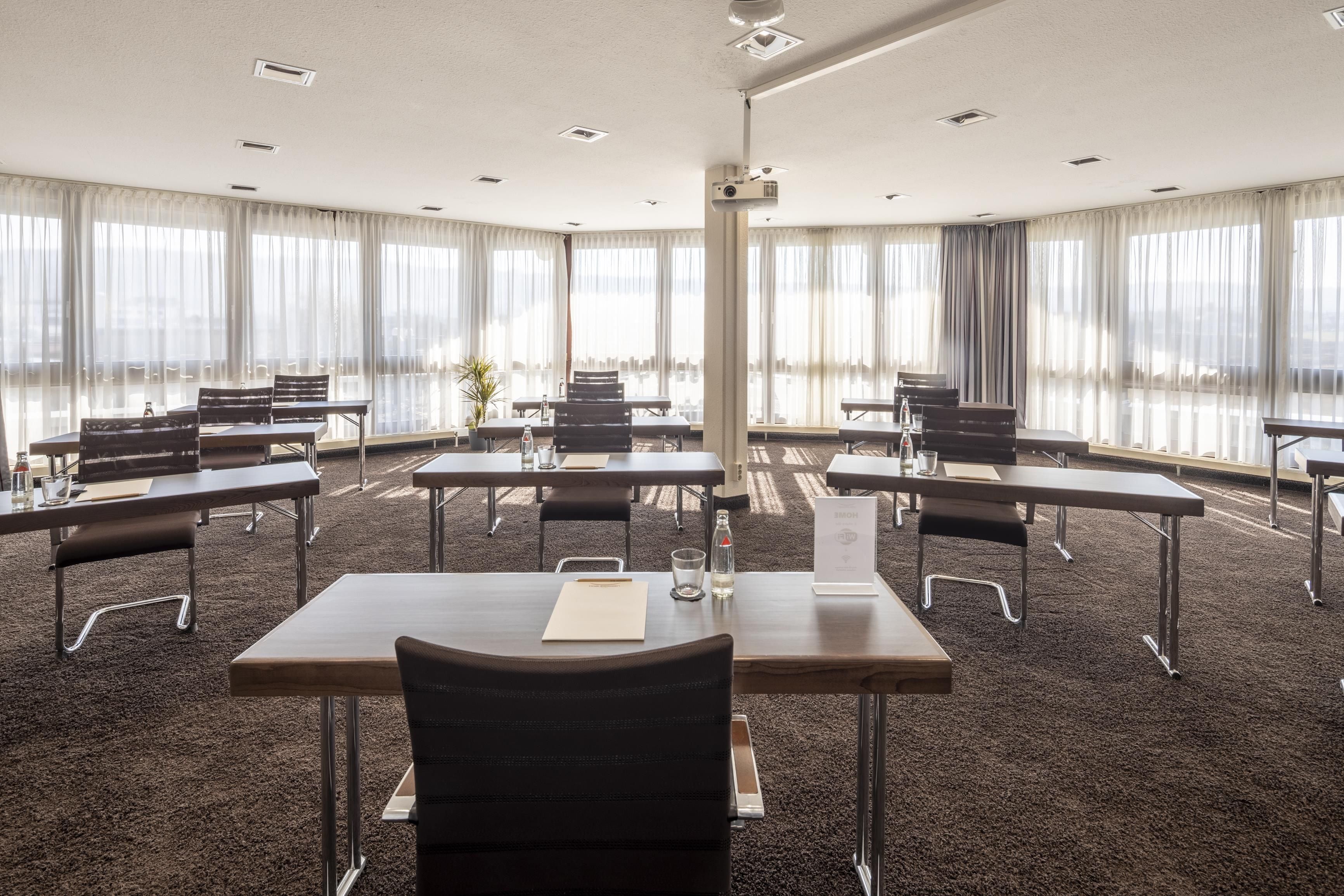 A conference room with multiple tables and chairs, large windows with sheer curtains, and a carpeted floor.