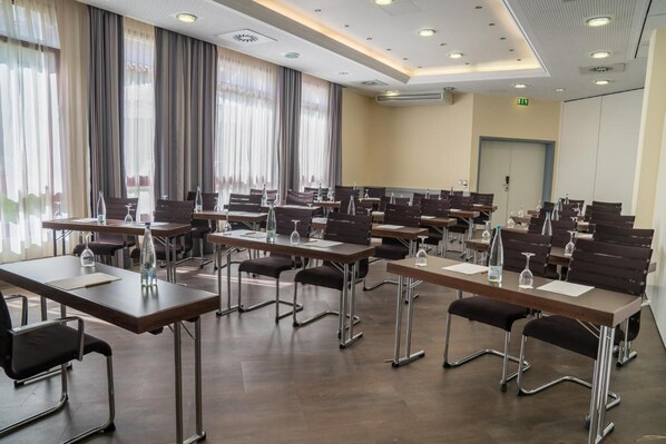 Un salón de conferencias con varias mesas y sillas, equipado con botellas de agua y vasos, ventanas grandes con cortinas y una unidad de aire acondicionado en el techo.
