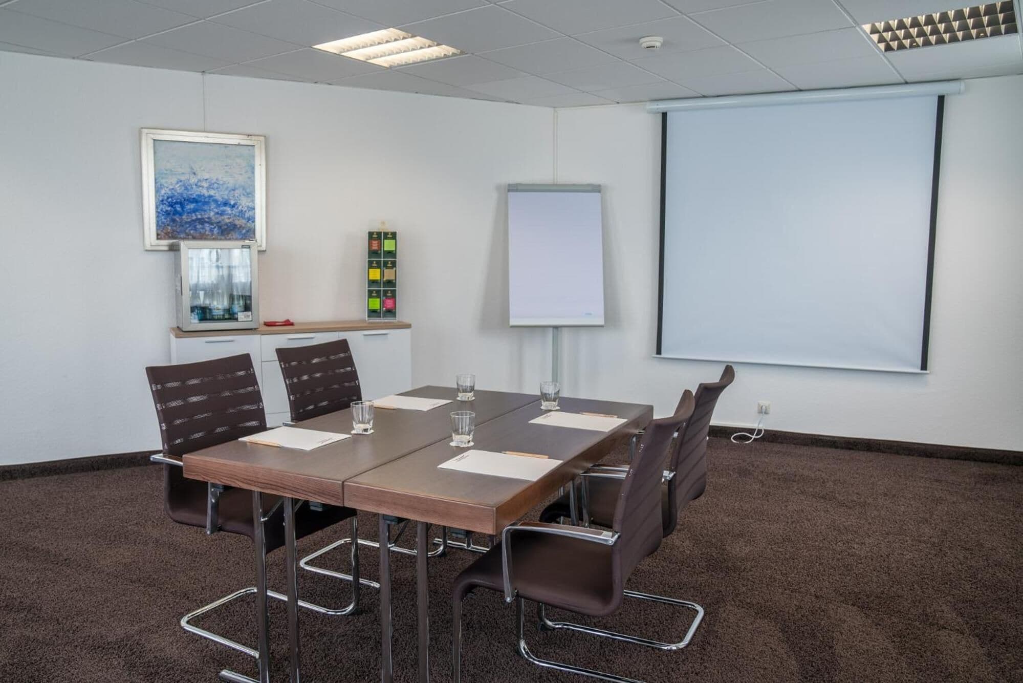 Ein Konferenzraum mit einem langen Tisch, sechs Stühlen, einer Projektionsleinwand, einer Tafel und einem Gemälde an der Wand.