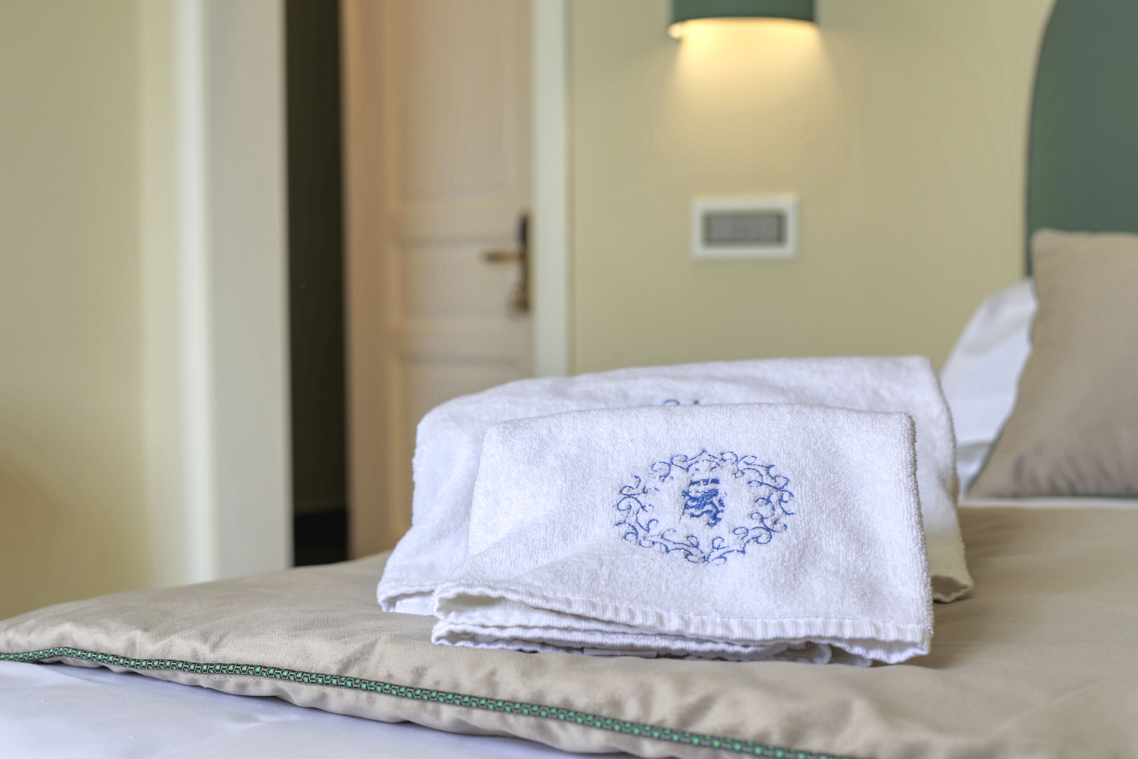 A neatly made bed with a folded towel and a pillow, a door in the background, and a wall-mounted light fixture.