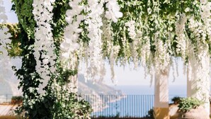A pergola with white flowers and greenery, overlooking a body of water. - Palazzo Confalone (Ravello)