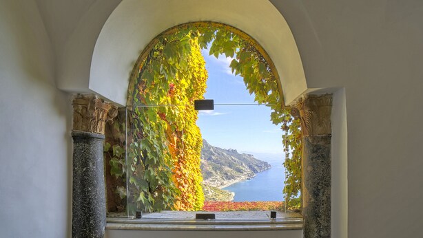 An arched window with a view of a coastal landscape, including a body of water and distant mountains. - Palazzo Confalone (Ravello)