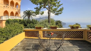A terrace with a table and chairs, potted plants, and a view of a building with arches and a mountain in the background. - Palazzo Confalone (Ravello)