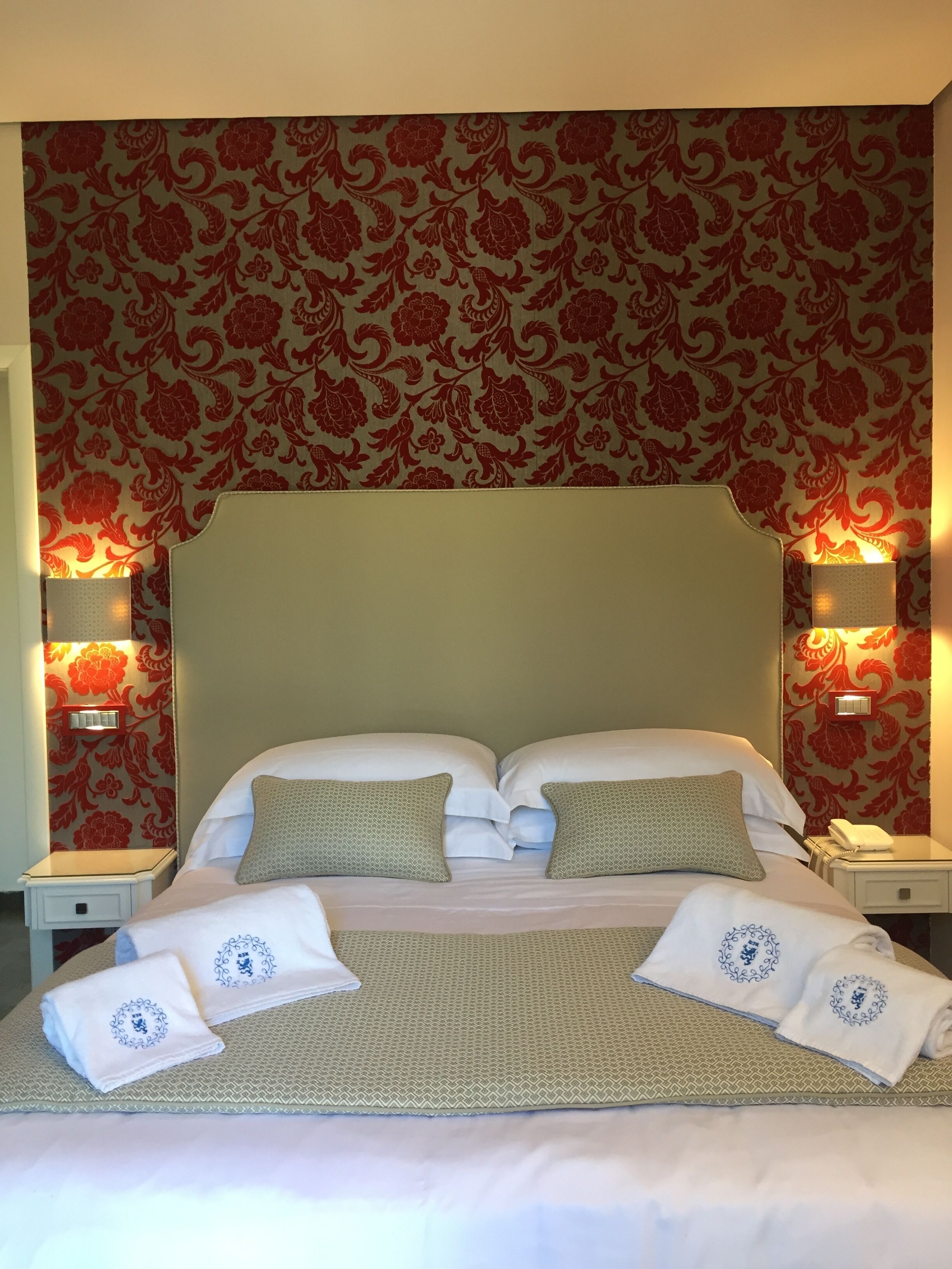 A double bed with white linens, two pillows, and a matching blanket. There are two bedside tables with lamps, a wall with a floral pattern, and a headboard.