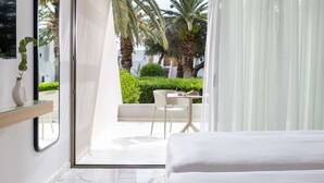A modern hotel room with a bed, a glass door leading to a balcony with a table and chairs, and a view of palm trees and greenery.