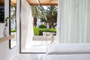 Una habitación de hotel moderna con una cama, un ventanal que da a un balcón con mesa y sillas, y vistas a palmeras y follaje.
