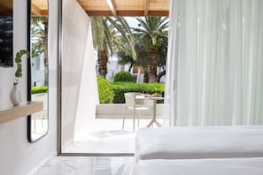 A modern hotel room with a bed, a glass door leading to a balcony with a table and chairs, and a view of palm trees and greenery.