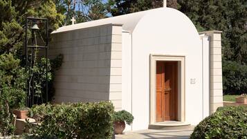 A small, white chapel with a wooden door, surrounded by greenery and a bell tower.