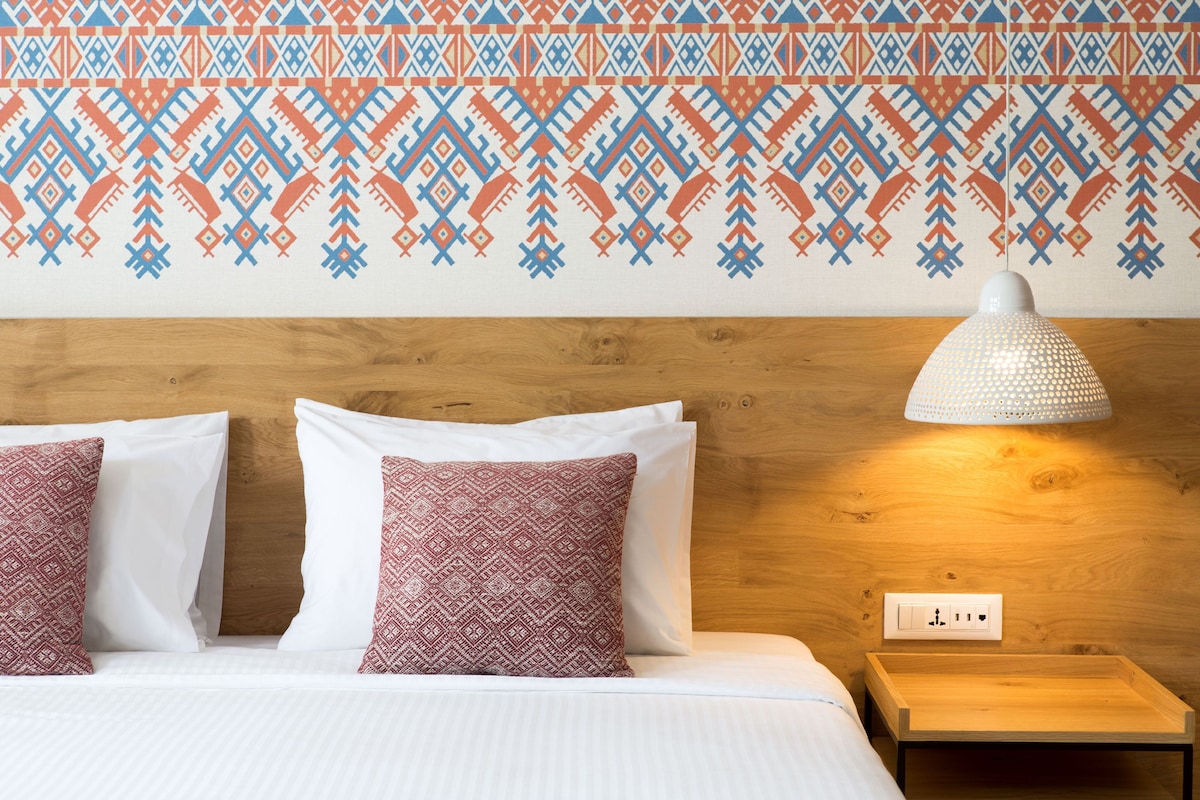 A bed with white linens and patterned pillows, a wooden headboard, a bedside lamp, and a wall-mounted power outlet.