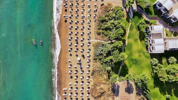 An aerial view of a beach with rows of umbrellas and beach chairs, a pathway leading to the water, and a building complex adjacent to the beach.