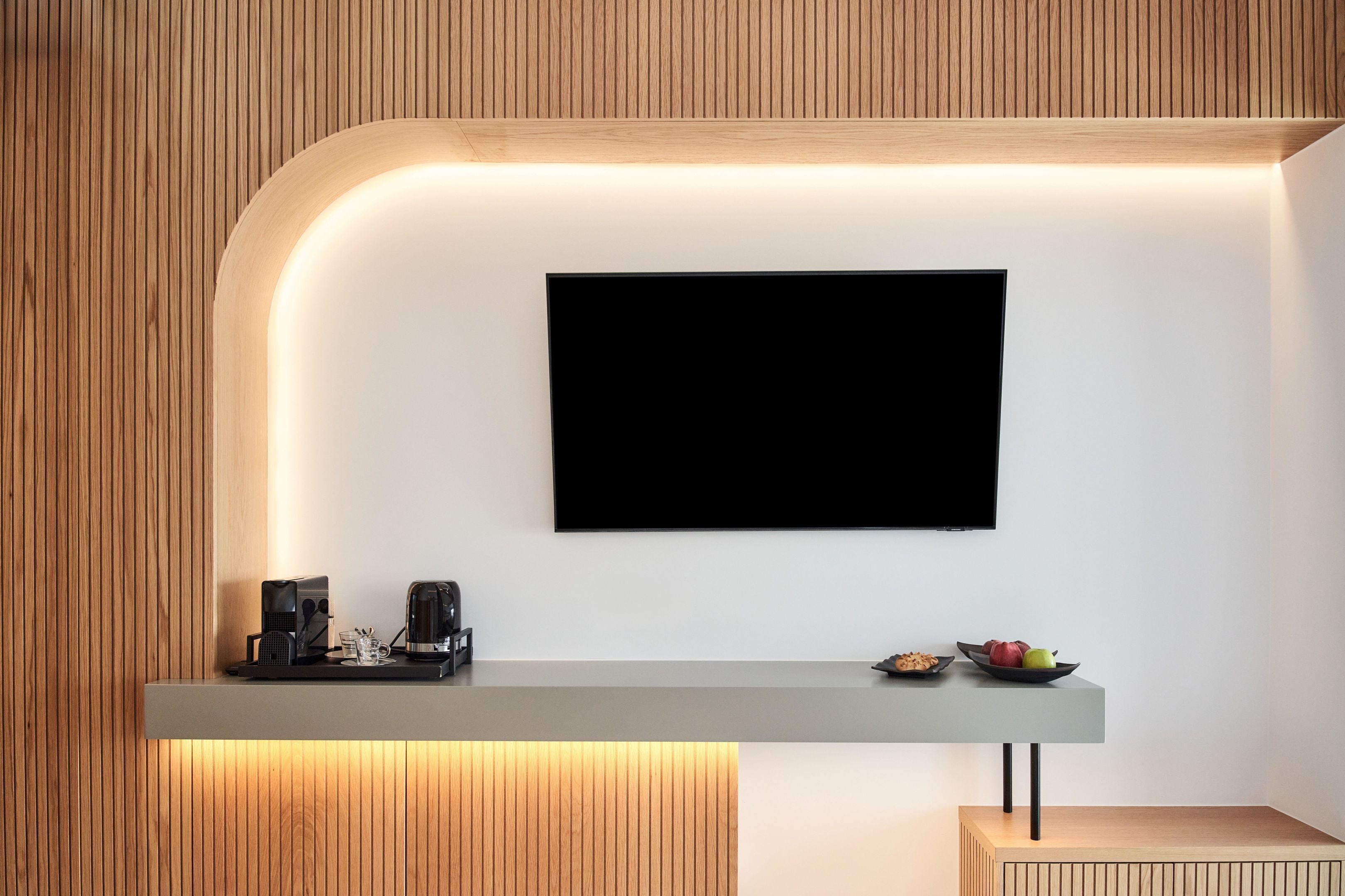 A modern reception area with a flat-screen TV mounted on a white wall, a reception desk with a coffee machine, and a small fruit bowl.