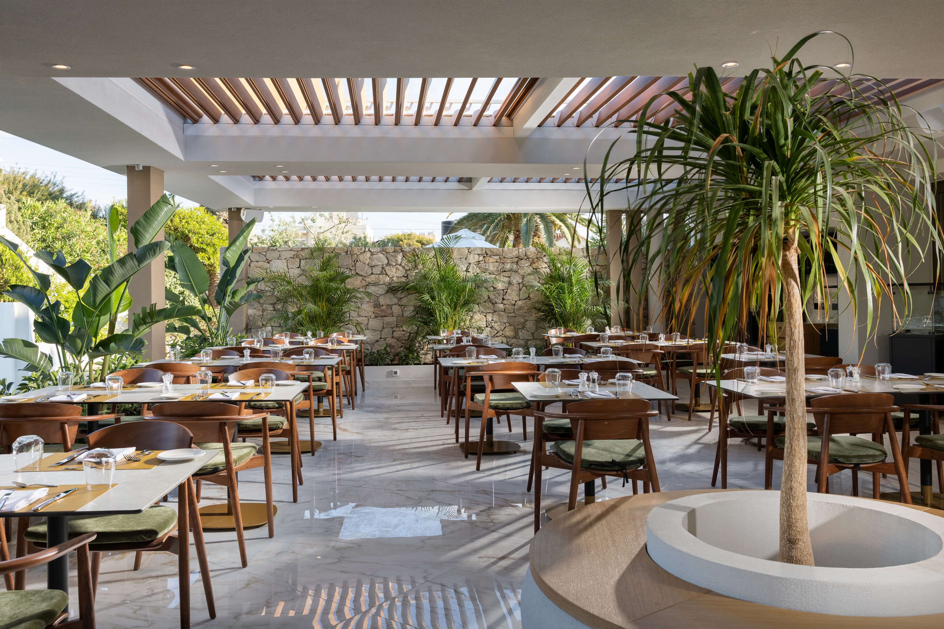 Un restaurante con terraza, paredes de piedra y un árbol central. Las mesas están dispuestas con manteles blancos y sillas verdes.