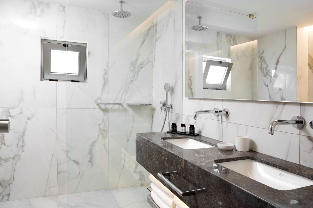 A modern bathroom with marble walls, two sinks, a large mirror, and a shower.