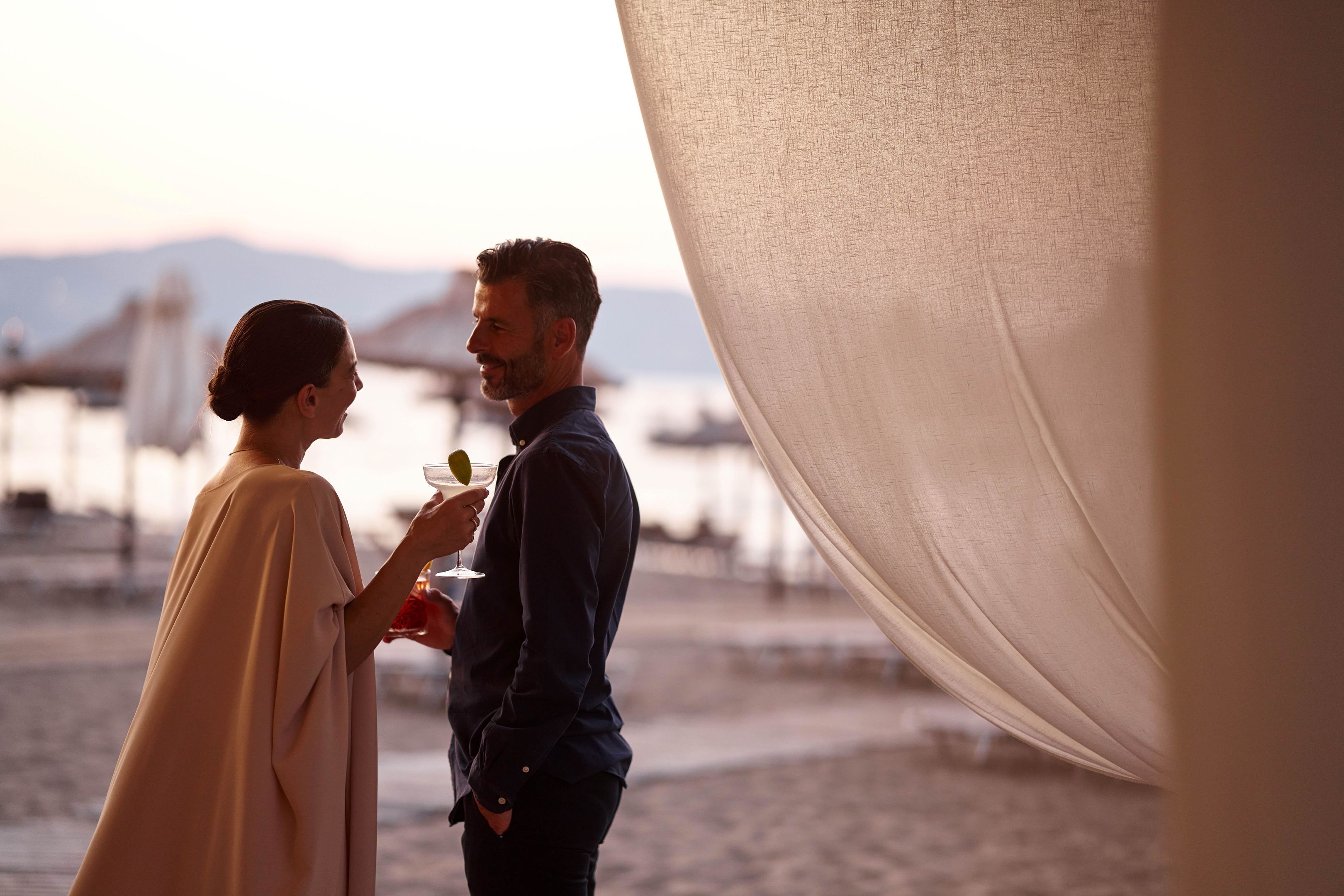 A couple standing on a beach with a backdrop of boats and a mountainous horizon.