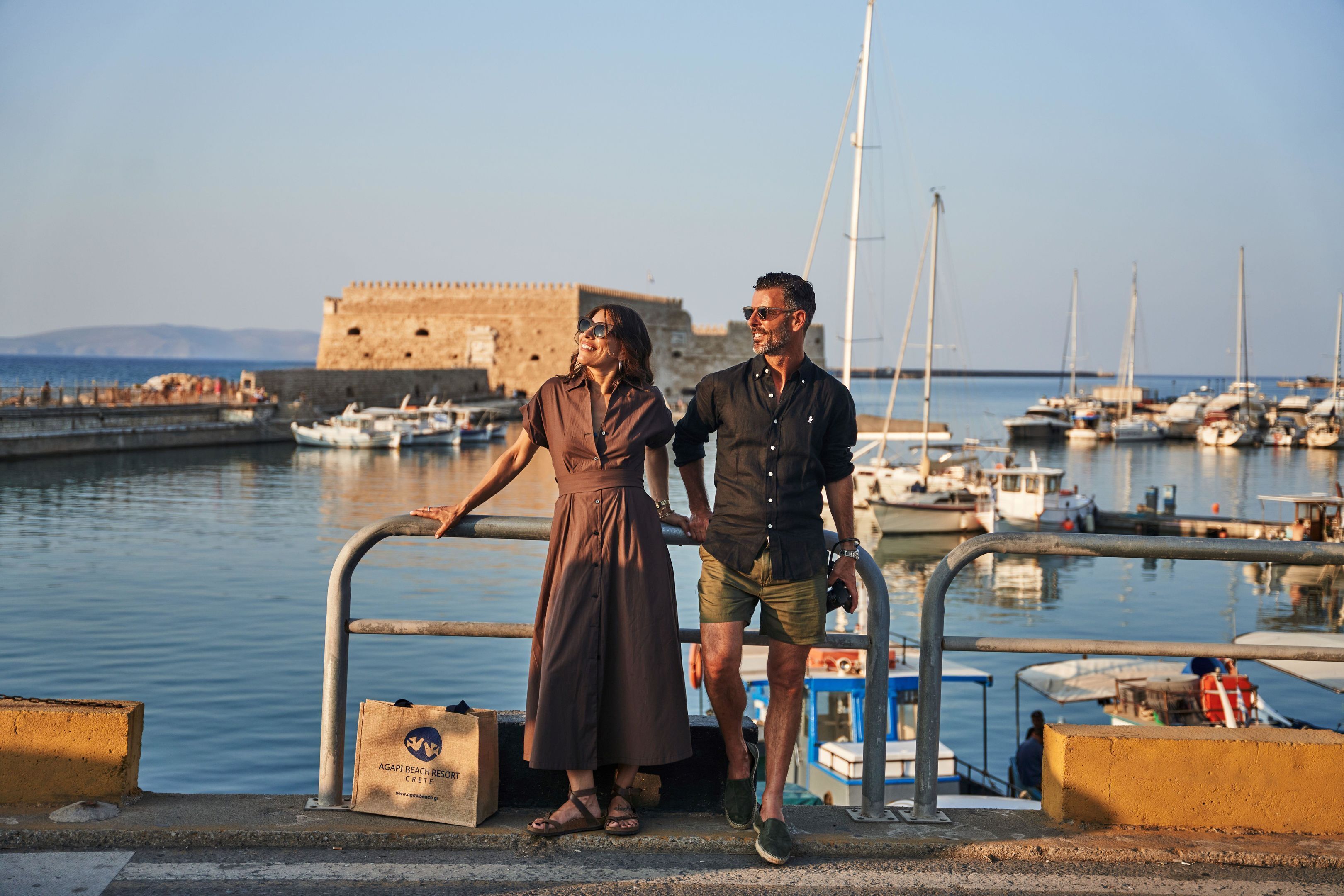 A couple standing by a waterfront with boats, a historic building, and a clear sky.