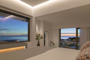 Una habitación de hotel moderna con una cama grande, una mesita de noche, una planta en maceta, un ventanal con vistas al mar y un balcón con barandilla.