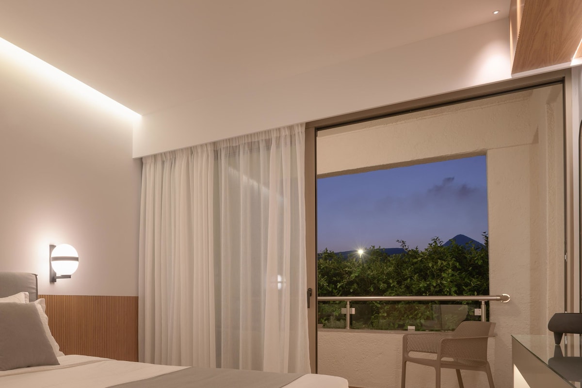 A modern hotel room with a large bed, a sitting area with a chair, and a balcony with a view of greenery and mountains.