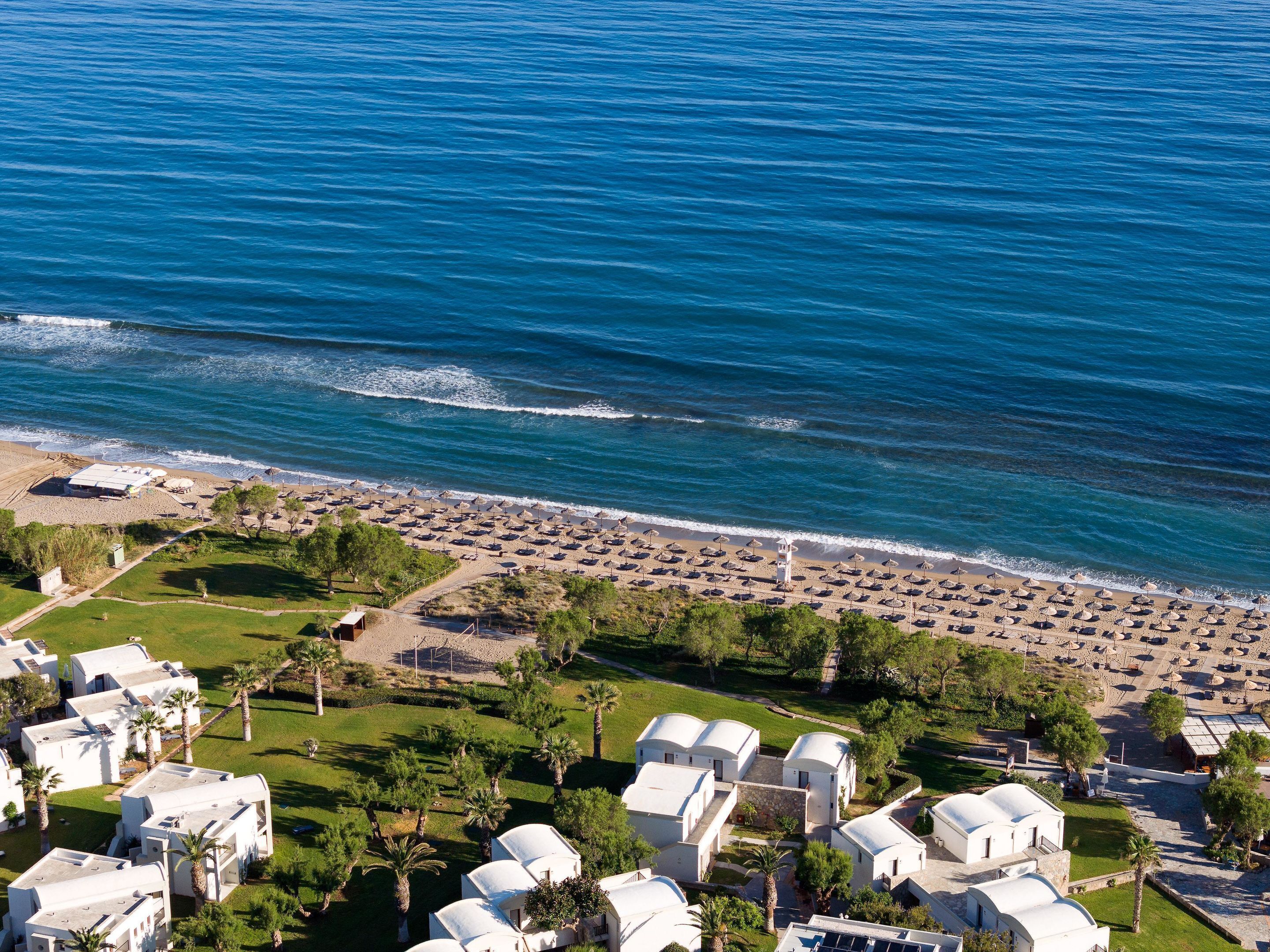 An aerial view of a coastal resort with beachside villas, a sandy beach with umbrellas, and a clear view of the ocean.