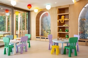 A children's play area with colorful chairs and tables, wooden shelves with toys, and murals on the walls.