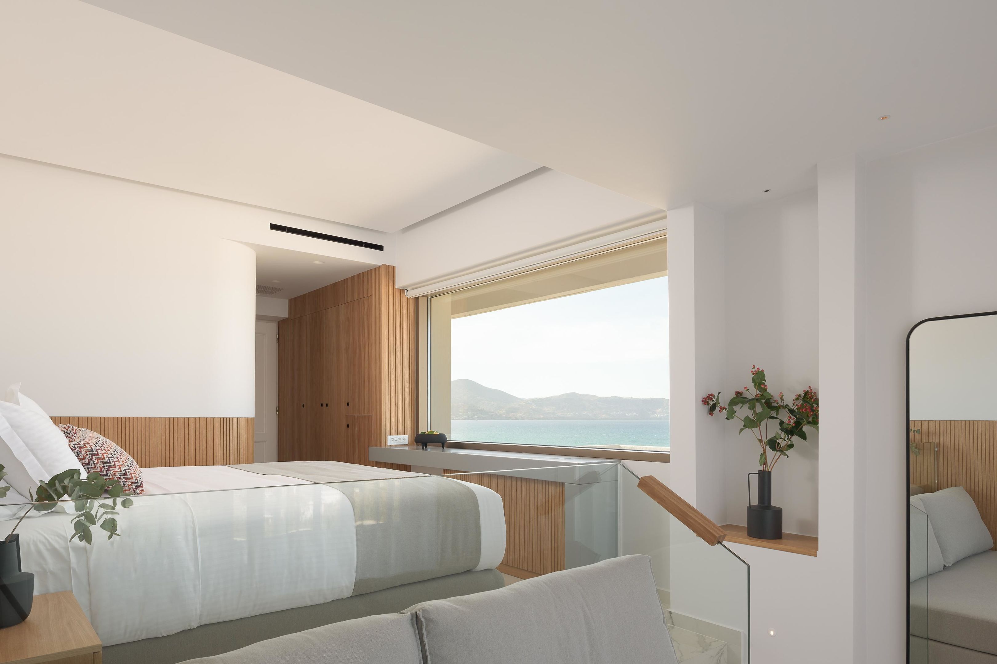 A modern bedroom with a large window, a bed, a bedside table, and a mirror.
