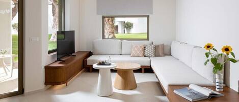 A modern living room with a sofa, a coffee table, a TV, and a window with a view of greenery.