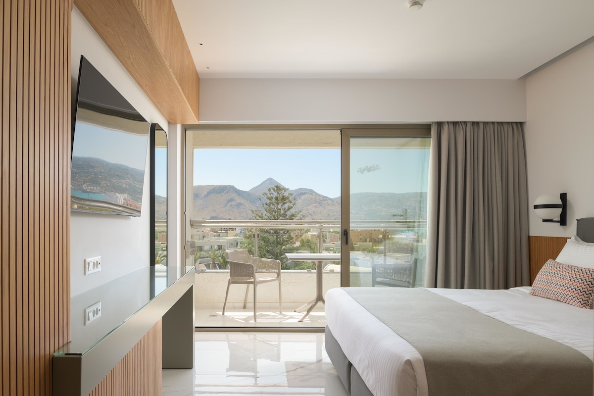A modern hotel room with a balcony, a bed, a TV, and a view of mountains.