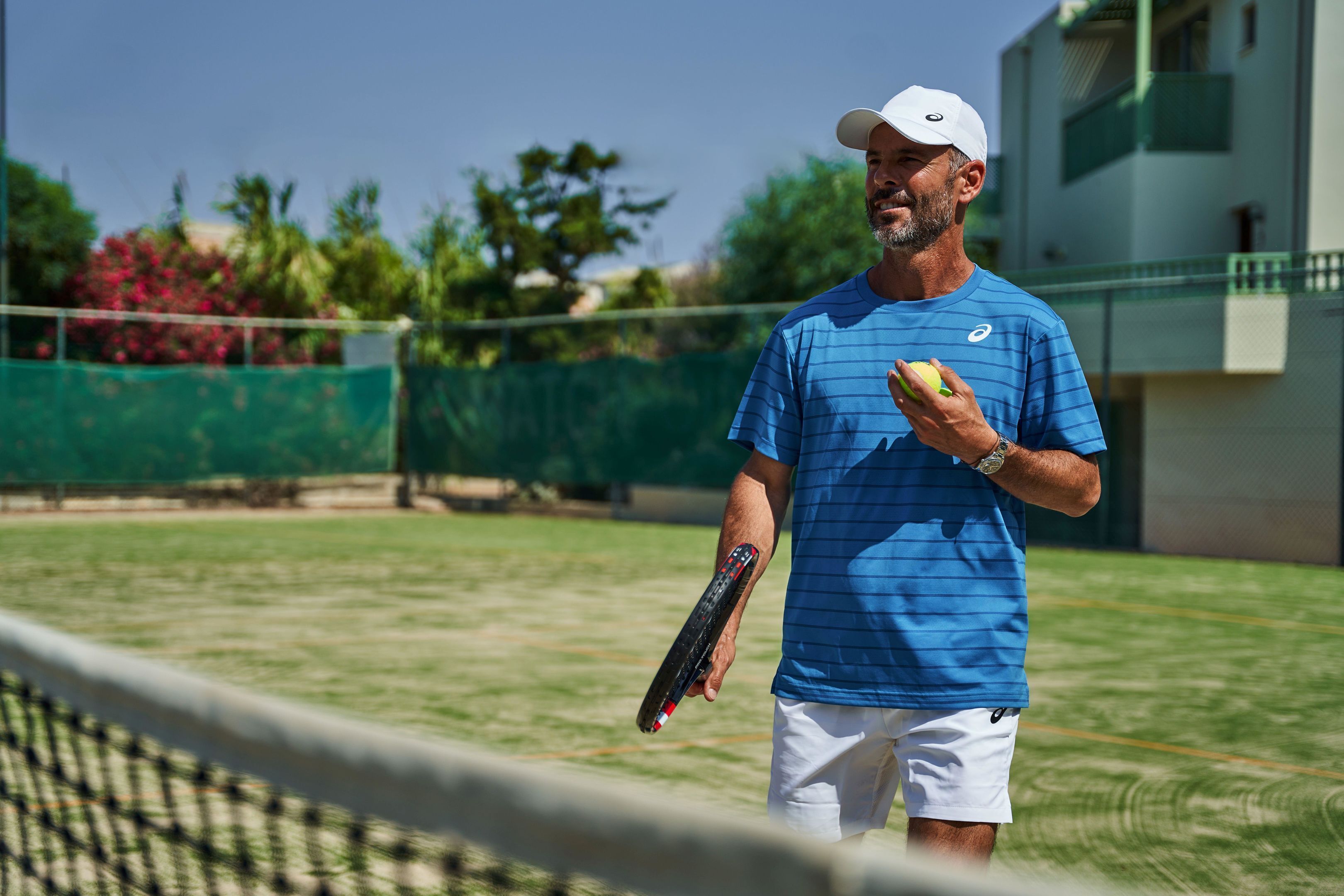 A tennis court with a net, a person holding a tennis ball and racket, and a building in the background.