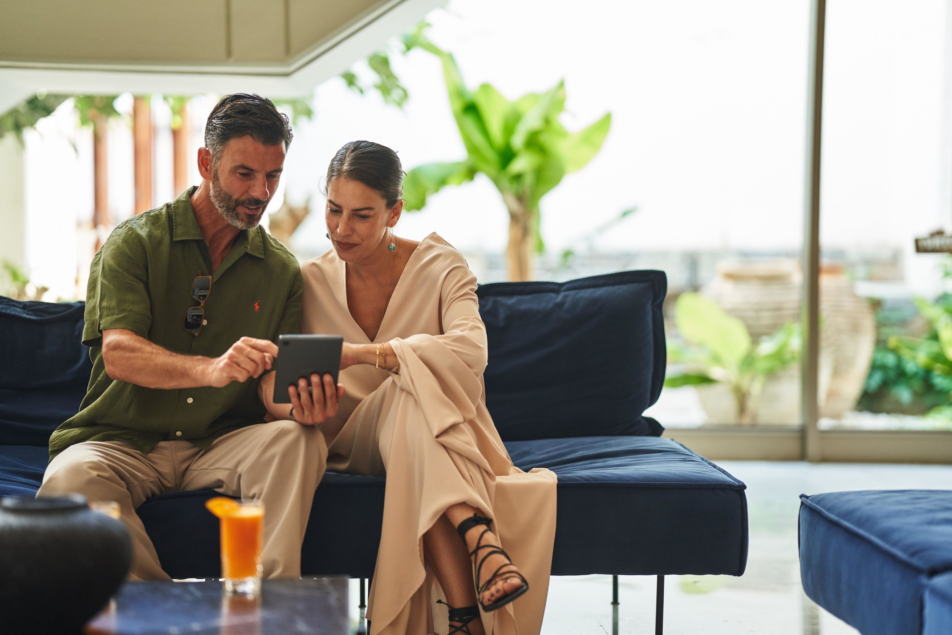 Un couple est assis sur un canapé bleu foncé, dans une pièce bien éclairée, avec une grande fenêtre, une plante et un verre de jus d’orange posé sur une table.
