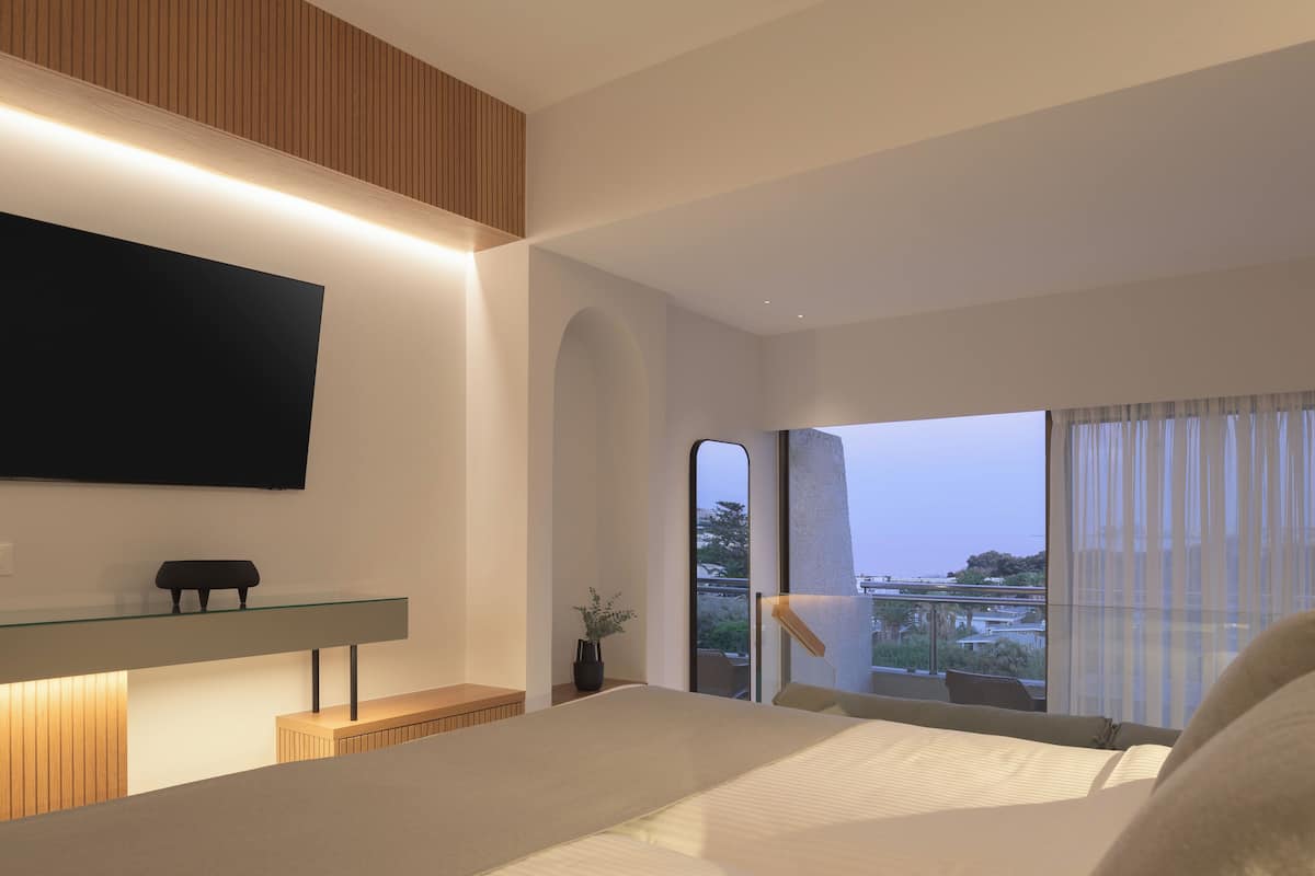 A modern hotel room with a large bed, a flat-screen TV mounted on the wall, a glass coffee table, and a view of the cityscape.