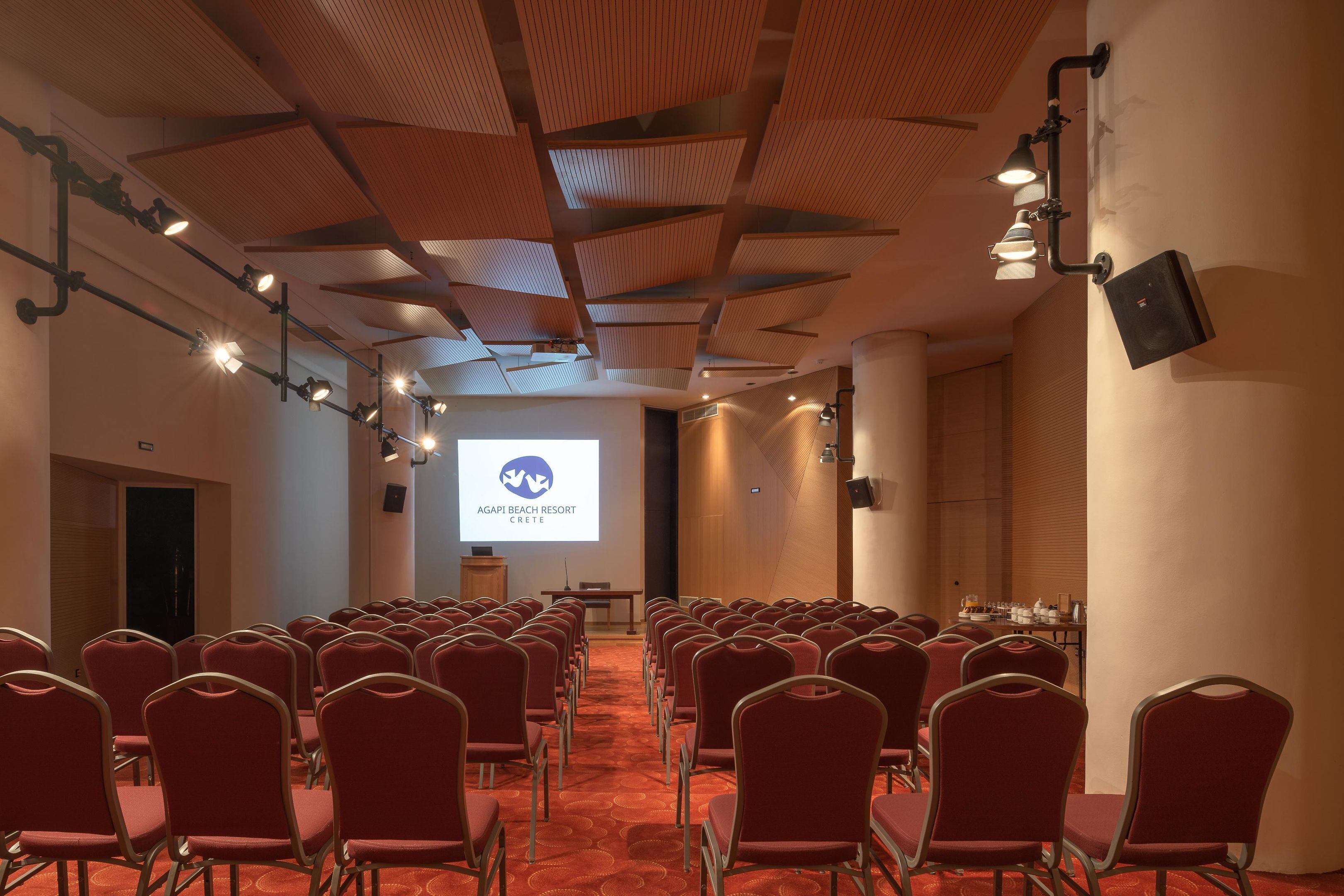 Ein Konferenzraum mit Reihen roter Stühle, einer Projektionsleinwand, die 'AGAPI BEACH RESORT CRETE' zeigt, und einer Holzbalkendecke.