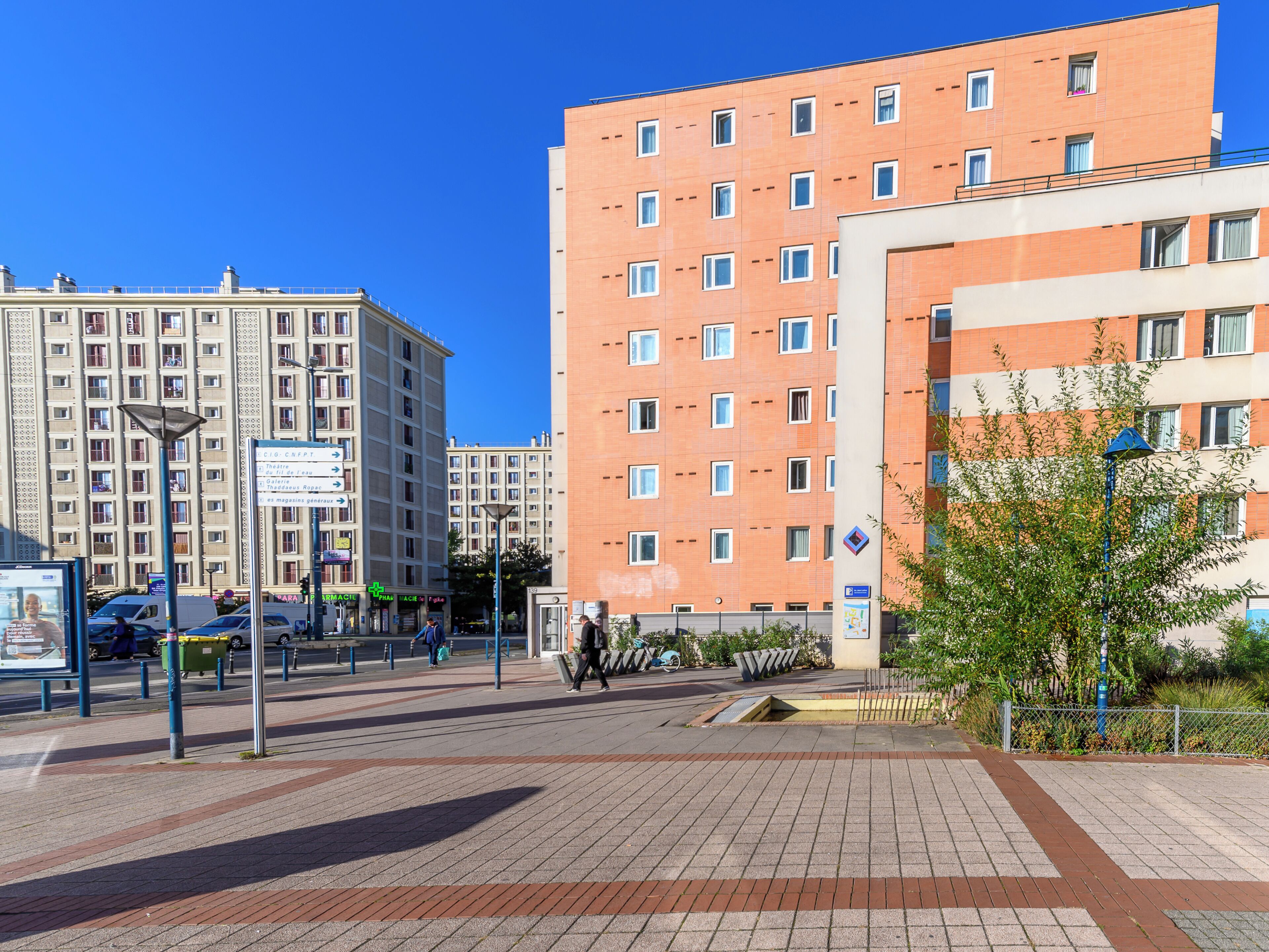 A modern urban square with a large red building, a smaller beige building, and a blue street lamp.
